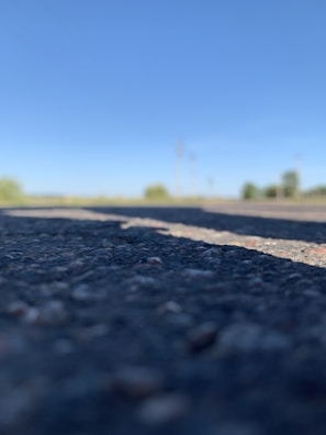 Close-up of an asphalt road with a clear blue sky in the background. The focus is on the textured surface of the road, while the background is blurred, showing vague shapes of trees or bushes along the horizon.