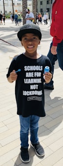 A young boy wearing a black cap and a black T-shirt with white text 'SCHOOLS ARE FOR LEARNING, NOT LOCKDOWN.' He is holding a bubble wand and bubble solution bottle. The background shows several people walking in an urban outdoor setting with palm trees and buildings.