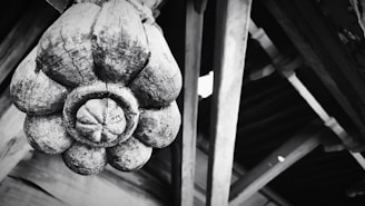 A large, intricately carved wooden ornament resembling a flower is attached to a wooden structure. The texture of the wood is rough and weathered, highlighting cracks and grain patterns. The background consists of wooden beams and planks, adding an architectural element.