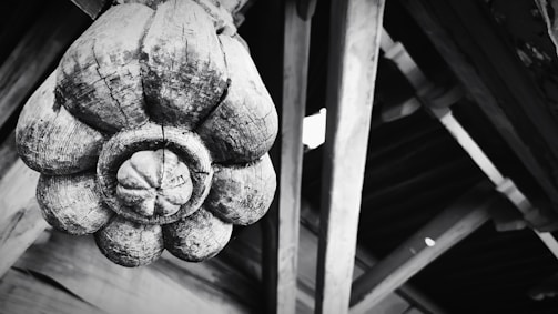 A large, intricately carved wooden ornament resembling a flower is attached to a wooden structure. The texture of the wood is rough and weathered, highlighting cracks and grain patterns. The background consists of wooden beams and planks, adding an architectural element.