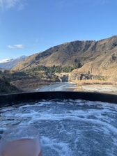 A serene mountainous landscape with clear blue skies and a river curving through the valley. In the foreground, there is a hot tub with bubbling water, and a glass of a cold beverage is visible.