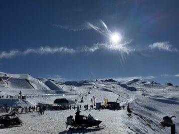 A snowy landscape with a ski resort is bustling with activity. People are skiing down the slopes, while others gather near the base. The sun shines brightly, creating a glistening effect on the snow. Ski lifts are transporting visitors up the mountain, and banners can be seen around the area. The scene is framed by clear skies and a few wispy clouds.
