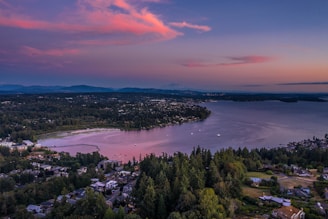 An aerial view captures a picturesque landscape featuring a body of water bordered by lush green forests and residential areas. The sky is painted with hues of pink and purple, suggesting a serene sunset or dawn. Rolling hills and distant mountains provide a tranquil backdrop.