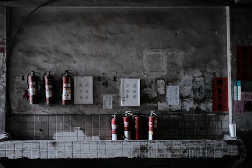 Several fire extinguishers are mounted on a weathered, industrial wall with various signs and papers attached above. The setting is dimly lit, giving it a slightly rustic and aged appearance. The lower part of the image shows more fire extinguishers placed on a countertop with visible wear.