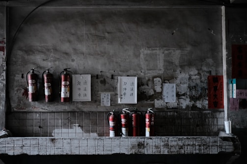 Several fire extinguishers are mounted on a weathered, industrial wall with various signs and papers attached above. The setting is dimly lit, giving it a slightly rustic and aged appearance. The lower part of the image shows more fire extinguishers placed on a countertop with visible wear.