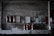 Several fire extinguishers are mounted on a weathered, industrial wall with various signs and papers attached above. The setting is dimly lit, giving it a slightly rustic and aged appearance. The lower part of the image shows more fire extinguishers placed on a countertop with visible wear.