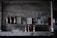 Several fire extinguishers are mounted on a weathered, industrial wall with various signs and papers attached above. The setting is dimly lit, giving it a slightly rustic and aged appearance. The lower part of the image shows more fire extinguishers placed on a countertop with visible wear.
