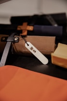 A close-up view shows an assortment of leather or fabric items, possibly bags or cases, featuring a brown and black strap. One item has a white tag with an engraved letter, indicating possible personalization. The items are arranged closely together on a dark surface.