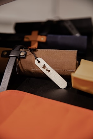 A close-up view shows an assortment of leather or fabric items, possibly bags or cases, featuring a brown and black strap. One item has a white tag with an engraved letter, indicating possible personalization. The items are arranged closely together on a dark surface.