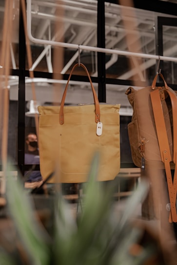 A stylish tan leather tote bag with brown handles hangs in a display, accompanied by a similar brown bag. The bags are elegantly showcased against a backdrop of a modern, industrial-style interior with visible black-framed windows and ceilings. Green plants partially obscure the view, adding an organic element to the scene.