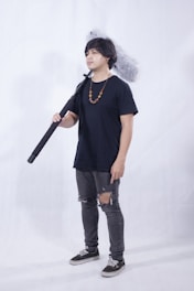 A person standing indoors holding a large boom microphone. They are wearing a navy blue t-shirt, jeans with rips at the knees, and black sneakers. A beaded necklace hangs around their neck. The background is plain and light-colored.
