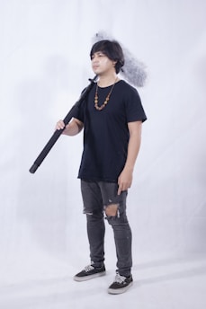A person standing indoors holding a large boom microphone. They are wearing a navy blue t-shirt, jeans with rips at the knees, and black sneakers. A beaded necklace hangs around their neck. The background is plain and light-colored.