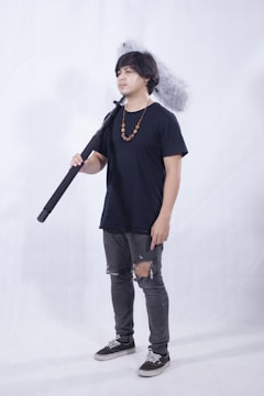 A person standing indoors holding a large boom microphone. They are wearing a navy blue t-shirt, jeans with rips at the knees, and black sneakers. A beaded necklace hangs around their neck. The background is plain and light-colored.