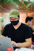 A person wearing a green cap, glasses, and a black face mask is using a laptop. They are seated indoors with another person in the background, also wearing a mask. Greenery and wood paneling are visible, suggesting a modern and calm setting.