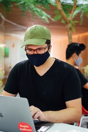 A person wearing a green cap, glasses, and a black face mask is using a laptop. They are seated indoors with another person in the background, also wearing a mask. Greenery and wood paneling are visible, suggesting a modern and calm setting.