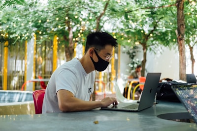 A person wearing a white t-shirt and a black face mask works on a laptop at an outdoor setting, surrounded by greenery and decorative lights. The ambiance includes tables and chairs with an additional person in the background.