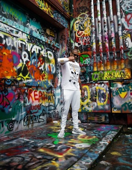 A person wearing white athletic clothing stands in front of a wall covered in vibrant and colorful graffiti. The individual is holding a gold chain and seems to be in a reflective pose. The surroundings are filled with various spray-painted words, drawings, and patterns, creating a dynamic and energetic atmosphere.