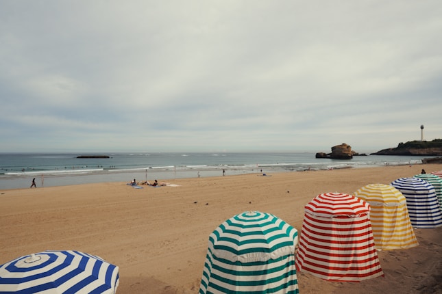 A sandy beach with a line of colorful striped beach tents in the foreground, set against a calm sea. A few people can be seen walking along the shore and some are swimming in the water. A rocky outcrop and a lighthouse are visible in the distance under a cloudy sky.