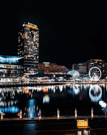 A nighttime cityscape featuring modern high-rise buildings with illuminated signs, including one labeled Sofitel. The scene is reflected in a calm body of water, showcasing bright lights and the illuminated outline of a Ferris wheel. The overall urban setting is vibrant, with visible architecture and street lamps.