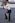 A person dressed in a security uniform is striking a playful pose, holding a blue baton in one hand and a notepad in the other. They are wearing sunglasses and black formal shoes, standing on a paved surface with a brick building and a parked car in the background.