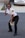A person dressed in a security uniform is striking a playful pose, holding a blue baton in one hand and a notepad in the other. They are wearing sunglasses and black formal shoes, standing on a paved surface with a brick building and a parked car in the background.