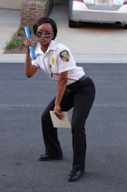 A person dressed in a security uniform is striking a playful pose, holding a blue baton in one hand and a notepad in the other. They are wearing sunglasses and black formal shoes, standing on a paved surface with a brick building and a parked car in the background.