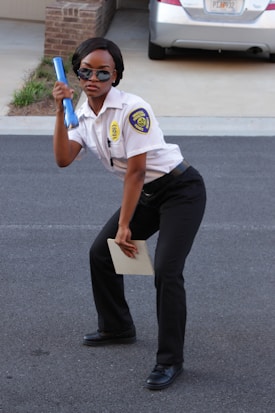 A person dressed in a security uniform is striking a playful pose, holding a blue baton in one hand and a notepad in the other. They are wearing sunglasses and black formal shoes, standing on a paved surface with a brick building and a parked car in the background.