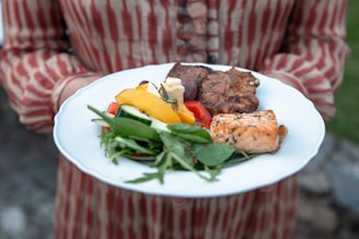 A person holding a white plate that contains a variety of grilled meats and fresh vegetables. The dish includes a piece of salmon, slices of grilled beef, mixed greens, and colorful bell pepper slices. The attire of the person features red and white striped fabric.