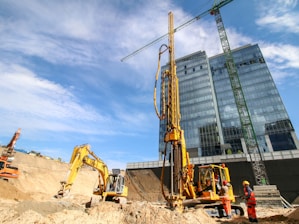 A construction site featuring two large yellow excavators and a worker in a hard hat and safety gear. The site is located in an urban environment with a modern high-rise glass building and a green crane in the background. The sky is clear with some scattered clouds, indicating a sunny day.