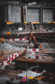 A construction site with heavy machinery, including an excavator, is actively operating next to a large, reinforced concrete structure. The area is surrounded by barriers and safety signs, and there are piles of materials and debris scattered around. A solitary worker is visible at the site amidst the equipment and construction materials.