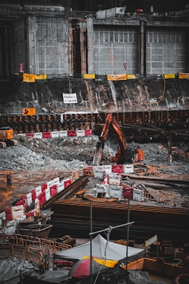 A construction site with heavy machinery, including an excavator, is actively operating next to a large, reinforced concrete structure. The area is surrounded by barriers and safety signs, and there are piles of materials and debris scattered around. A solitary worker is visible at the site amidst the equipment and construction materials.