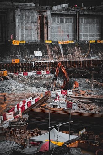 A construction site with heavy machinery, including an excavator, is actively operating next to a large, reinforced concrete structure. The area is surrounded by barriers and safety signs, and there are piles of materials and debris scattered around. A solitary worker is visible at the site amidst the equipment and construction materials.
