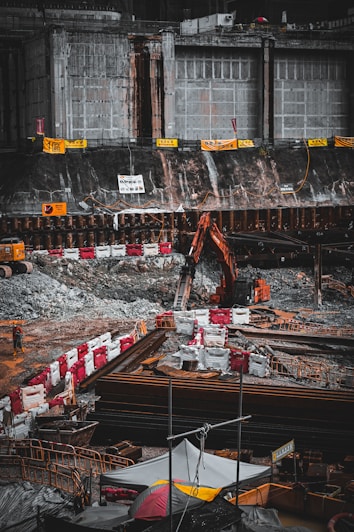 A construction site with heavy machinery, including an excavator, is actively operating next to a large, reinforced concrete structure. The area is surrounded by barriers and safety signs, and there are piles of materials and debris scattered around. A solitary worker is visible at the site amidst the equipment and construction materials.