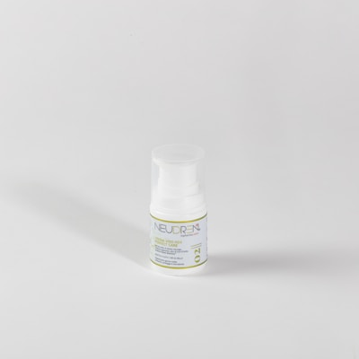 A small white plastic bottle with a pump dispenser sits against a plain light gray background. The bottle features a label with green and blue text, including the word 'NEUDREN'. The lighting creates a soft shadow to the left.