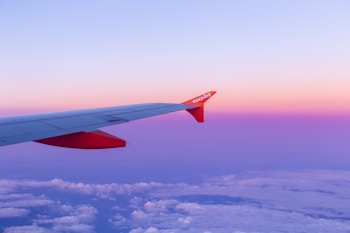 An airplane wing with a red tip and an easyJet logo extends over a landscape of fluffy clouds. The sky is a gradient of pink and purple as the sun sets or rises.