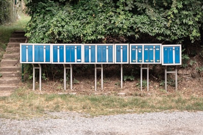A row of blue mailboxes is mounted on metal stands in an outdoor setting, surrounded by green foliage. The mailboxes are of uniform size and have numbers and postal logos on them. They are positioned on a patch of grass and gravel, with a staircase visible in the background leading up to a pathway.