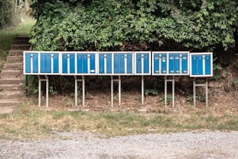 A row of blue mailboxes is mounted on metal stands in an outdoor setting, surrounded by green foliage. The mailboxes are of uniform size and have numbers and postal logos on them. They are positioned on a patch of grass and gravel, with a staircase visible in the background leading up to a pathway.