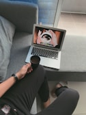 A person is sitting on a gray couch, holding a cup of coffee. They are watching an anime on a laptop placed on the couch next to them. The screen displays hands holding a swirling object. The person is casually dressed in dark clothing and a watch, with slippers visible on their feet. The setting appears comfortable and relaxed, with natural light coming from a window.