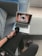 A person is sitting on a gray couch, holding a cup of coffee. They are watching an anime on a laptop placed on the couch next to them. The screen displays hands holding a swirling object. The person is casually dressed in dark clothing and a watch, with slippers visible on their feet. The setting appears comfortable and relaxed, with natural light coming from a window.