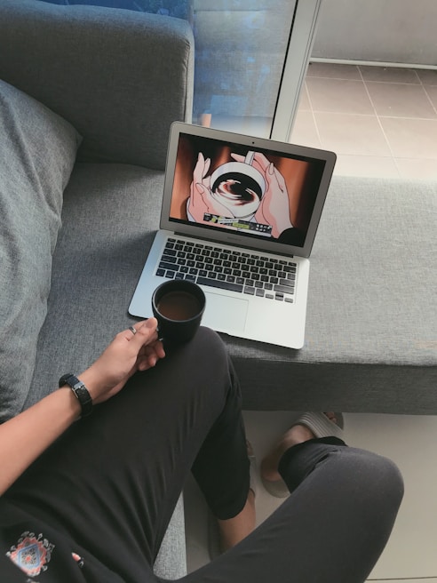 A person is sitting on a gray couch, holding a cup of coffee. They are watching an anime on a laptop placed on the couch next to them. The screen displays hands holding a swirling object. The person is casually dressed in dark clothing and a watch, with slippers visible on their feet. The setting appears comfortable and relaxed, with natural light coming from a window.