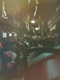 Inside a dimly lit bus, passengers are seated, some facing forward while others are facing backward. The interior features overhead lights and grab handles hanging from the ceiling. The atmosphere is quiet, suggesting a nighttime or late evening setting.