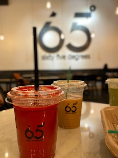 Three plastic cups with drinks are placed on a marble countertop. The focus is on the foreground cup, which contains a vivid red drink. Two cups in the background hold light brown and green drinks. The cups have straws and are branded with the text '65 degrees'. The background is slightly blurred, featuring a large wall sign that repeats the '65 degrees' branding.