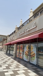 A restaurant storefront with Arabic and English signage reading 'Shake Shack' on the exterior wall. The building features architectural elements like decorative columns and a patterned tile walkway. Large posters on the windows display images of burgers and promotional text in Arabic.