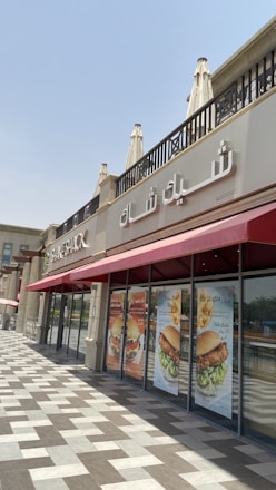 A restaurant storefront with Arabic and English signage reading 'Shake Shack' on the exterior wall. The building features architectural elements like decorative columns and a patterned tile walkway. Large posters on the windows display images of burgers and promotional text in Arabic.
