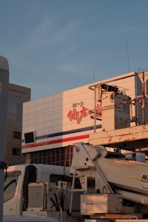 A large building facade with text in red and blue lines, with an industrial vehicle parked in front. The vehicle appears to be a construction or utility truck with machinery attached.
