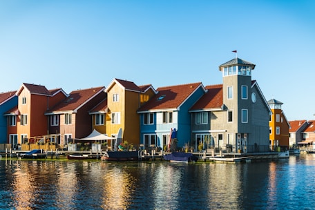 Colorful waterfront houses with red roofs and various painted facades are reflected in the calm water. The buildings have a distinctive Scandinavian architectural style, with large windows and wooden panels. A small boat is docked in front of the houses, and the clear blue sky adds to the serene atmosphere.