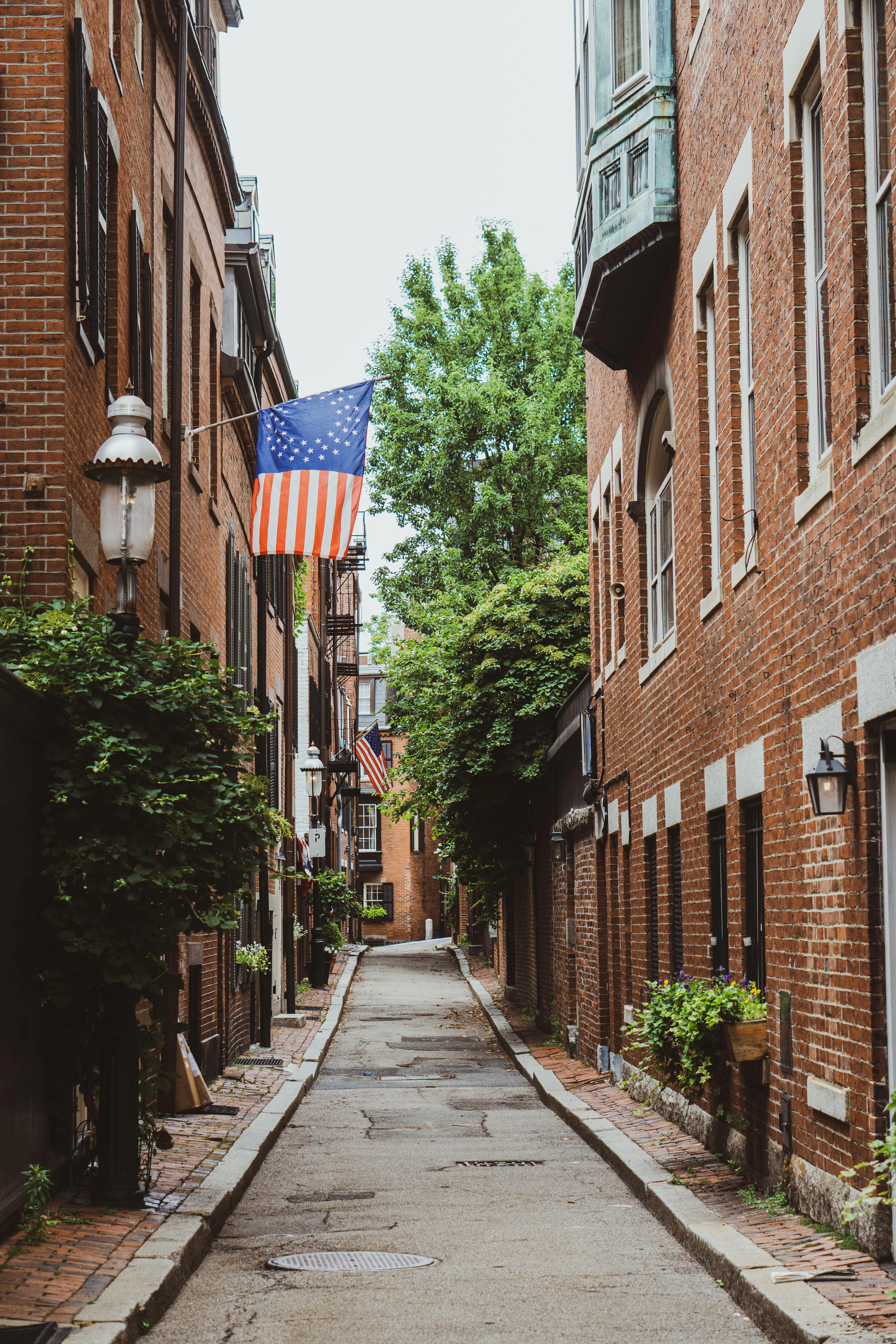 A narrow brick backstreet on Beacon Hill in Boston, empty in soft light, with a thirteen-star colonial flag and an iron gas lantern hanging out over the pavement.