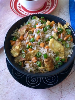 A bowl of fried rice featuring pieces of scrambled eggs, diced carrots, and green peas. The rice is served in a black bowl with a decorative swirl pattern at the base. A smaller bowl sits on a red and white plate in the background, partially visible.