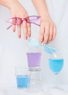 Two hands are holding sunglasses over three glasses filled with colorful liquid. The left hand holds pink sunglasses, and the right hand holds blue sunglasses. The liquids in the glasses are light blue and purple, with one glass being an inverted clear cup.