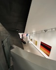 A modern art gallery with a sleek, angular architectural design. The interior features large, brightly colored abstract paintings on the walls. A solitary figure walks through the exhibition space, which is illuminated by overhead track lighting, highlighting the artwork on display.